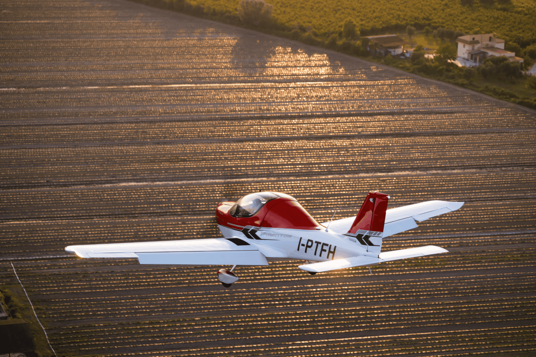 Small red airplane flying over farmland at sunset.
