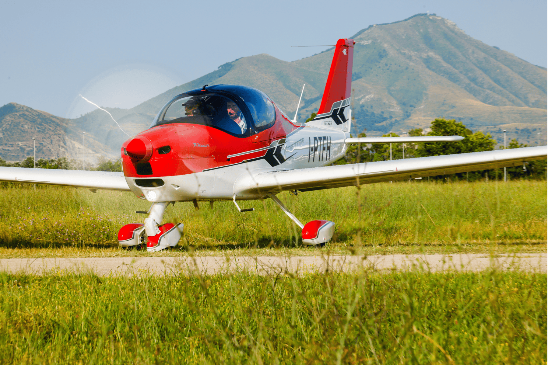 Red and white plane on grassy field with mountains.